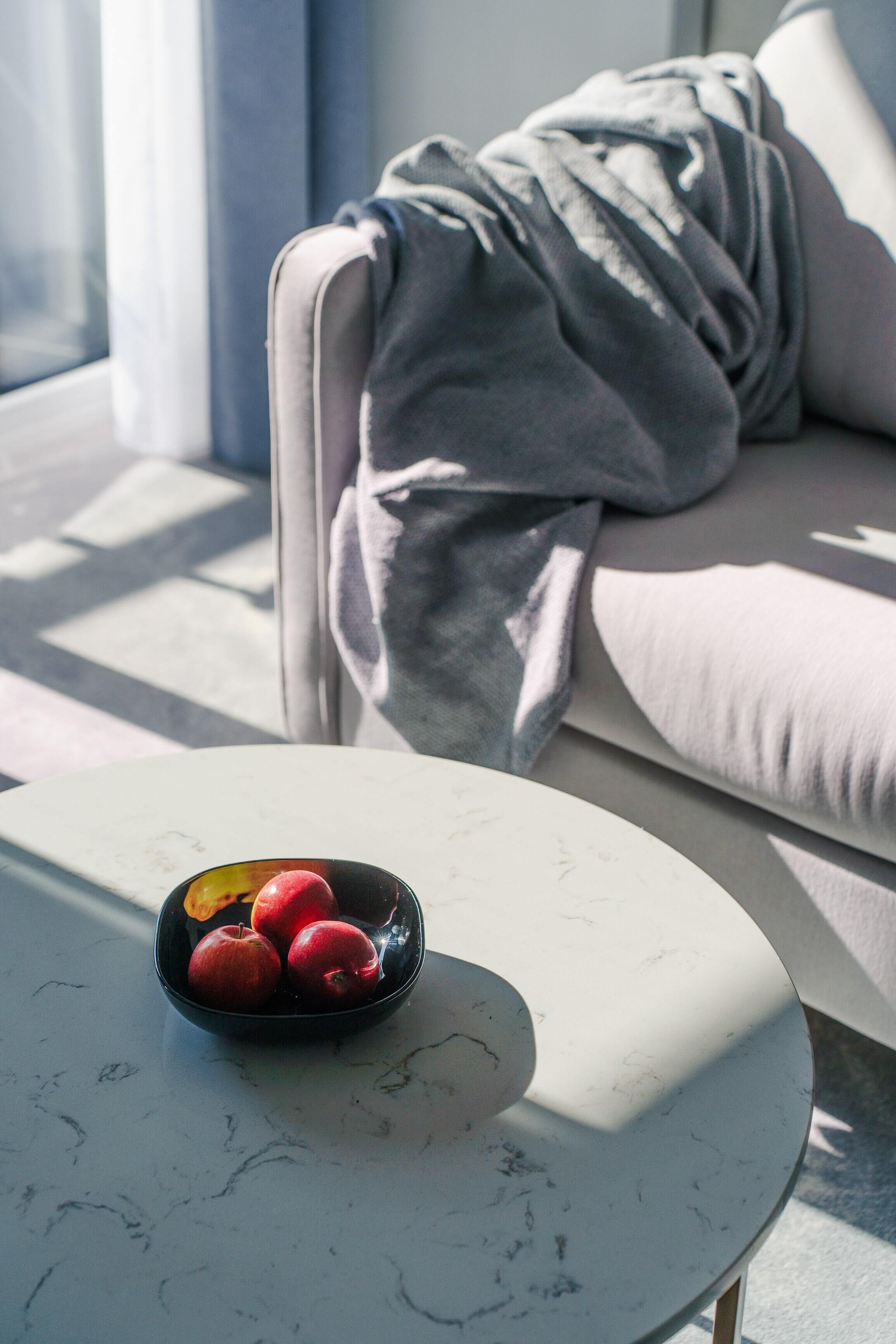 Una sala de estar moderna con una mesa redonda blanca, un bol de manzanas y un sofá gris con una manta.