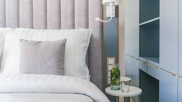 Een net opgemaakt bed met kussens, een nachtkastje met een glas en een plant, en een lamp aan de muur.