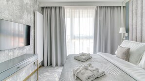 Une chambre d’hôtel moderne dotée d’un grand téléviseur à écran plat, d’un lit bien fait avec deux serviettes, et d’une grande fenêtre ornée de rideaux légers.