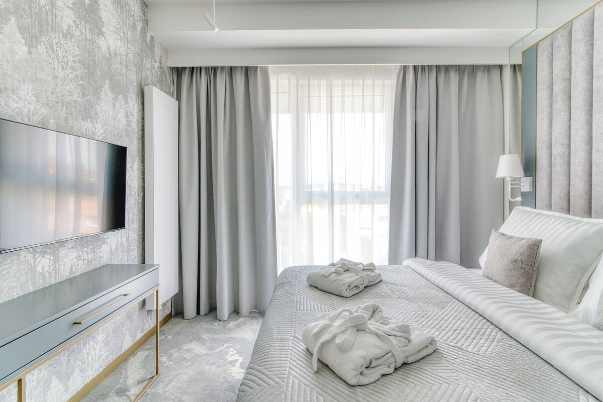 Una habitación de hotel moderna con un televisor de pantalla plana grande, una cama bien hecha con dos toallas, y un ventanal con cortinas traslúcidas.