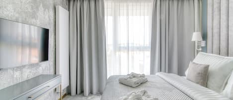A modern hotel room with a large flat-screen TV, a neatly made bed with two towels, and a large window with sheer curtains.