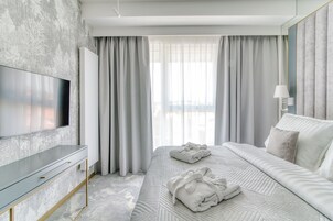 Una habitación de hotel moderna con un televisor de pantalla plana grande, una cama bien hecha con dos toallas, y un ventanal con cortinas traslúcidas.