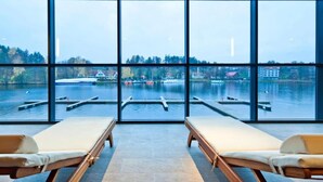 Une pièce dotée de grandes fenêtres donnant sur un lac, de deux chaises longues et offrant une vue sur les bateaux et les bâtiments situés de l’autre côté de l’eau.