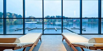 Una habitación con grandes ventanales que dan a un lago, dos tumbonas y vistas a barcos y edificios al otro lado del agua.