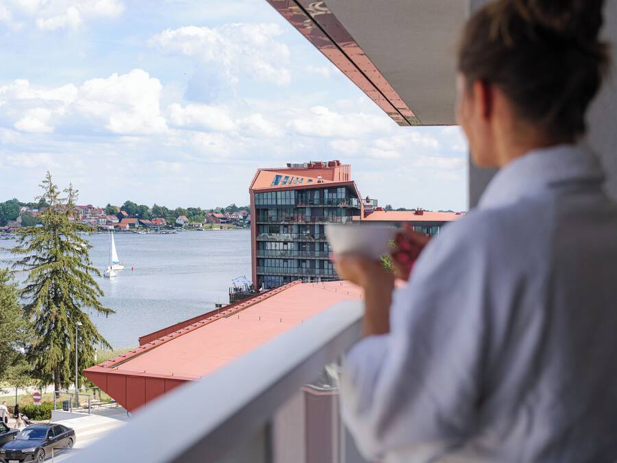 Un balcón con vista a un lago, un velero y un edificio moderno al otro lado del agua.