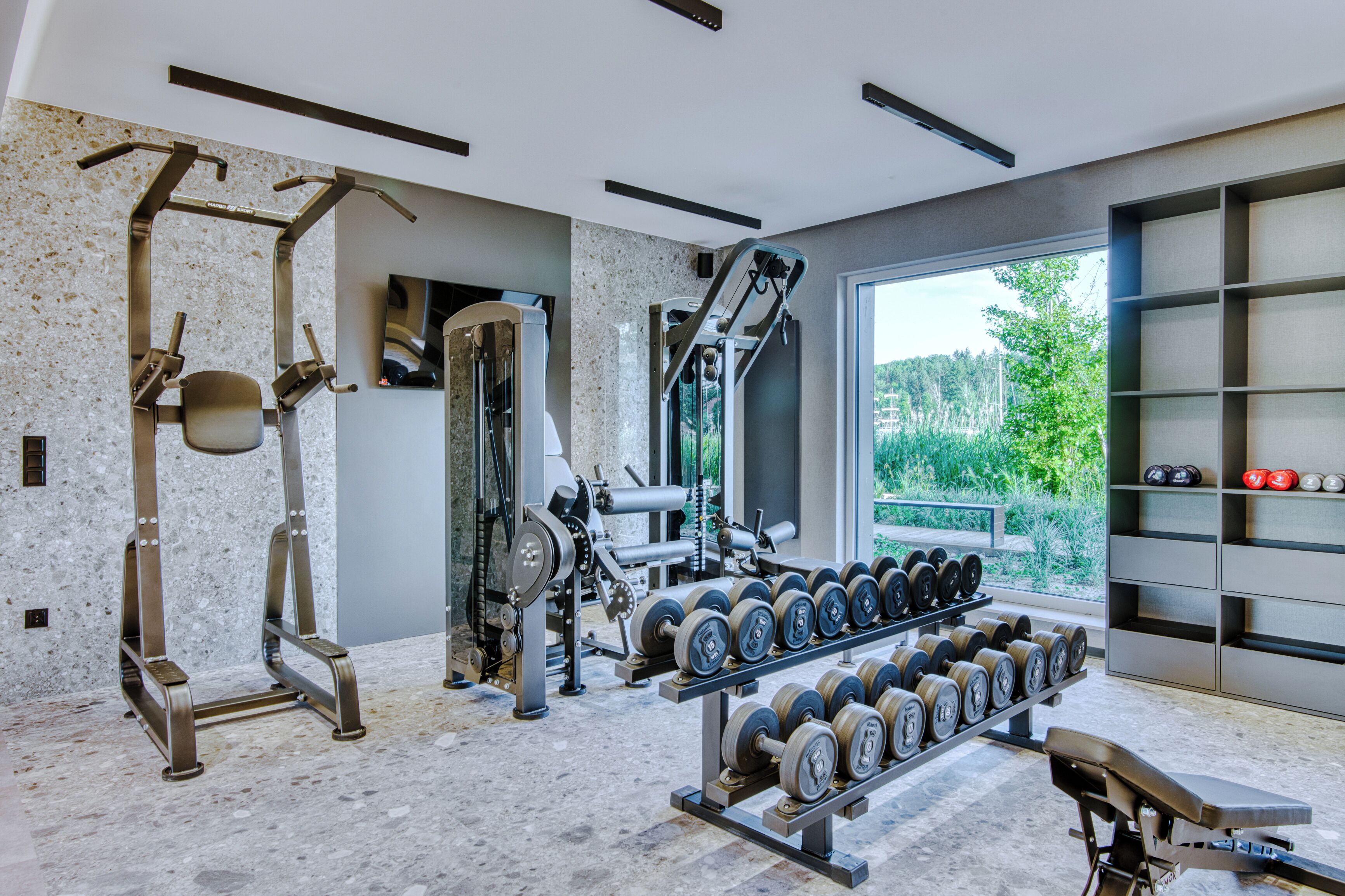 Academia moderna com diversos equipamentos de ginástica, um banco e uma grande janela com vista para vegetação.