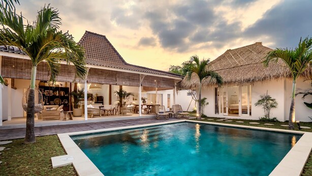 A poolside area with a clear pool, a covered outdoor seating area, and a thatched-roof building.