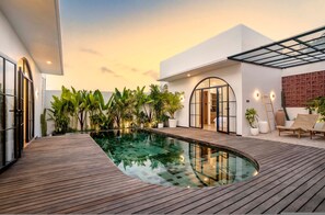 Ein moderner Poolbereich im Freien mit Holzdecking, umgeben von üppiger Bepflanzung und einer gläsernen Struktur.