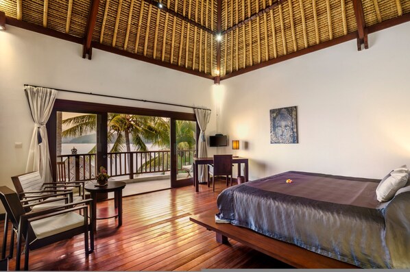 Una habitación con cama, escritorio y silla. Tiene un balcón con vistas a palmeras y al océano.