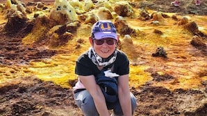 A person crouching on a rocky terrain with yellowish formations, another person in the background, and a clear sky.