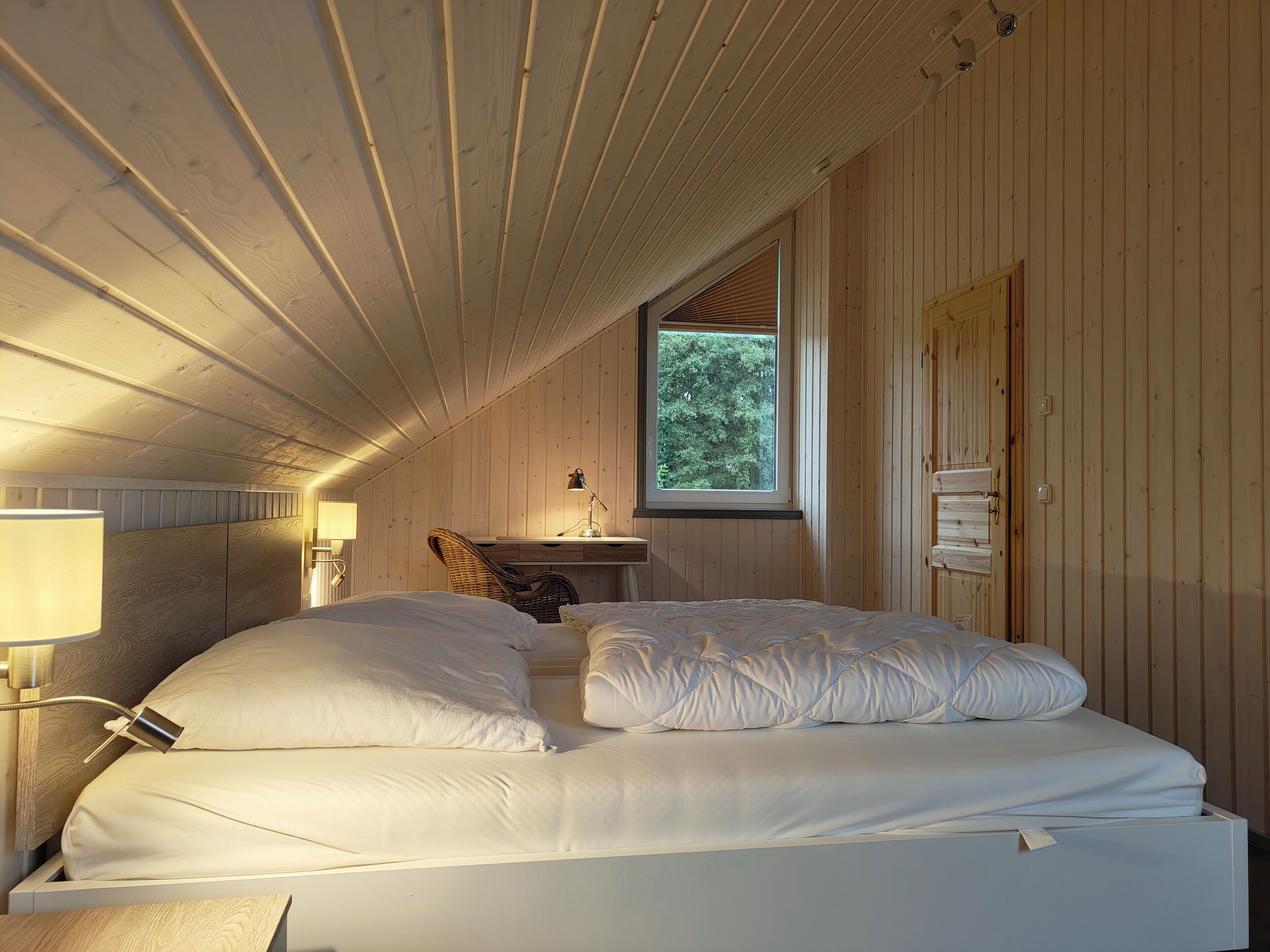 Een gezellige slaapkamer met een houten plafond, een groot bed met witte lakens, een bureau met lamp en een raam met uitzicht op bomen.