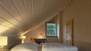 Tunnelmallinen makuuhuone, jossa on puinen katto, suuri sänky valkoisilla vuodevaatteilla, työpöytä lampun kanssa ja ikkuna, josta avautuu näkymä puihin.