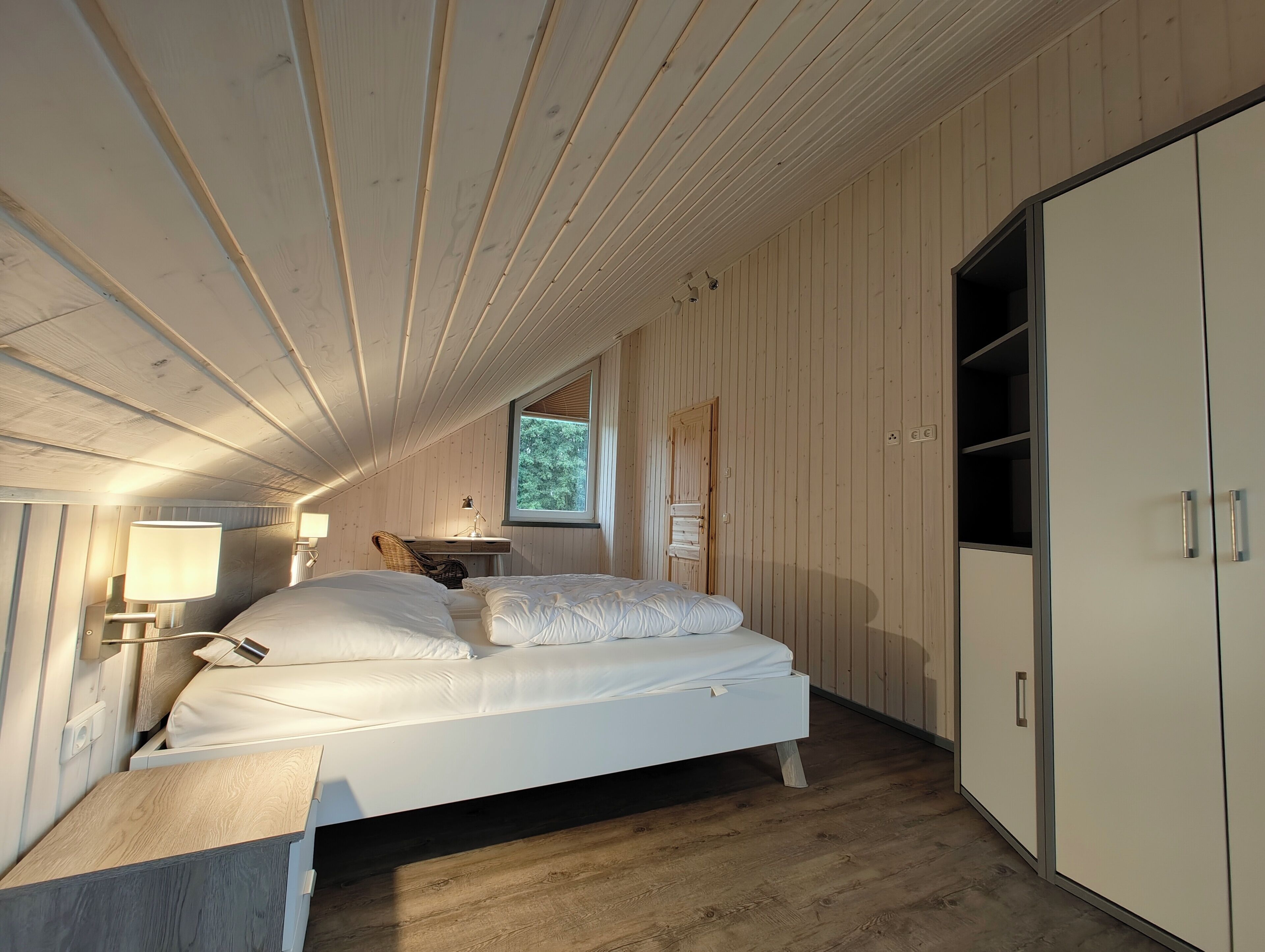 A bedroom with a wooden ceiling, a bed with white bedding, a desk with a chair, a window with a view of trees, and a wardrobe.