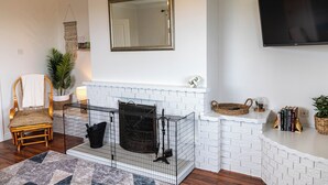 Ein Wohnzimmer mit Kamin aus weißem Mauerwerk, einem großen Spiegel, einem Fernseher, einem Korbstuhl und einem Bücherregal.