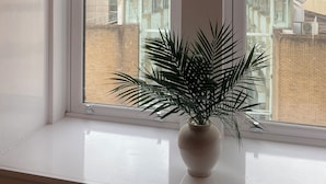 A window with a white roller blind, a potted plant on the windowsill, and a view of buildings outside. - Central Paisley Apartment- Airport/glasgow Access (Paisley)