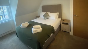 A neatly made bed with a green blanket and white towels, a bedside table with a remote control, and a window with blinds. - Central Paisley Apartment- Airport/glasgow Access (Paisley)