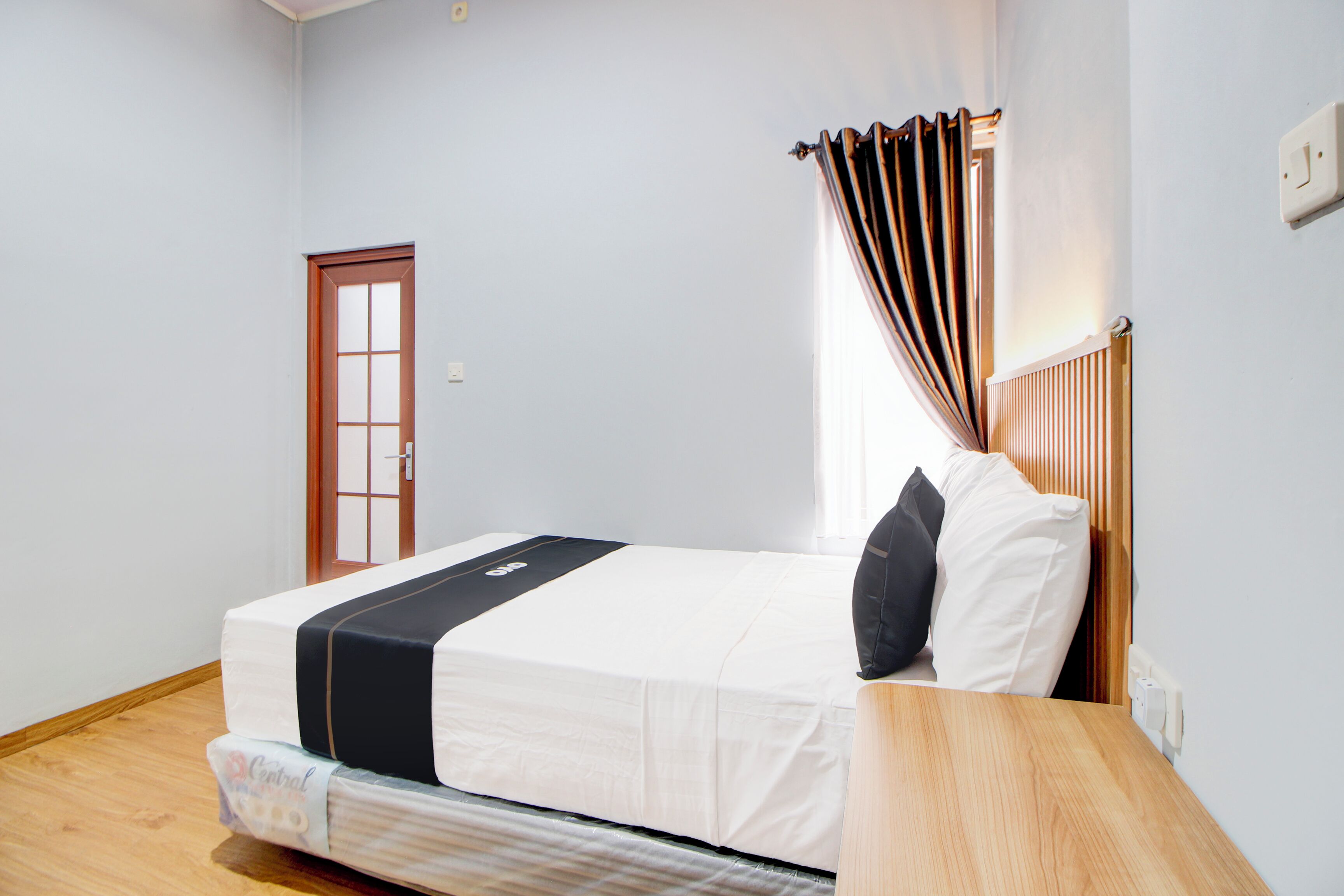 Uma cama bem arrumada com colcha preta e branca, cabeceira de madeira e uma janela com cortinas.