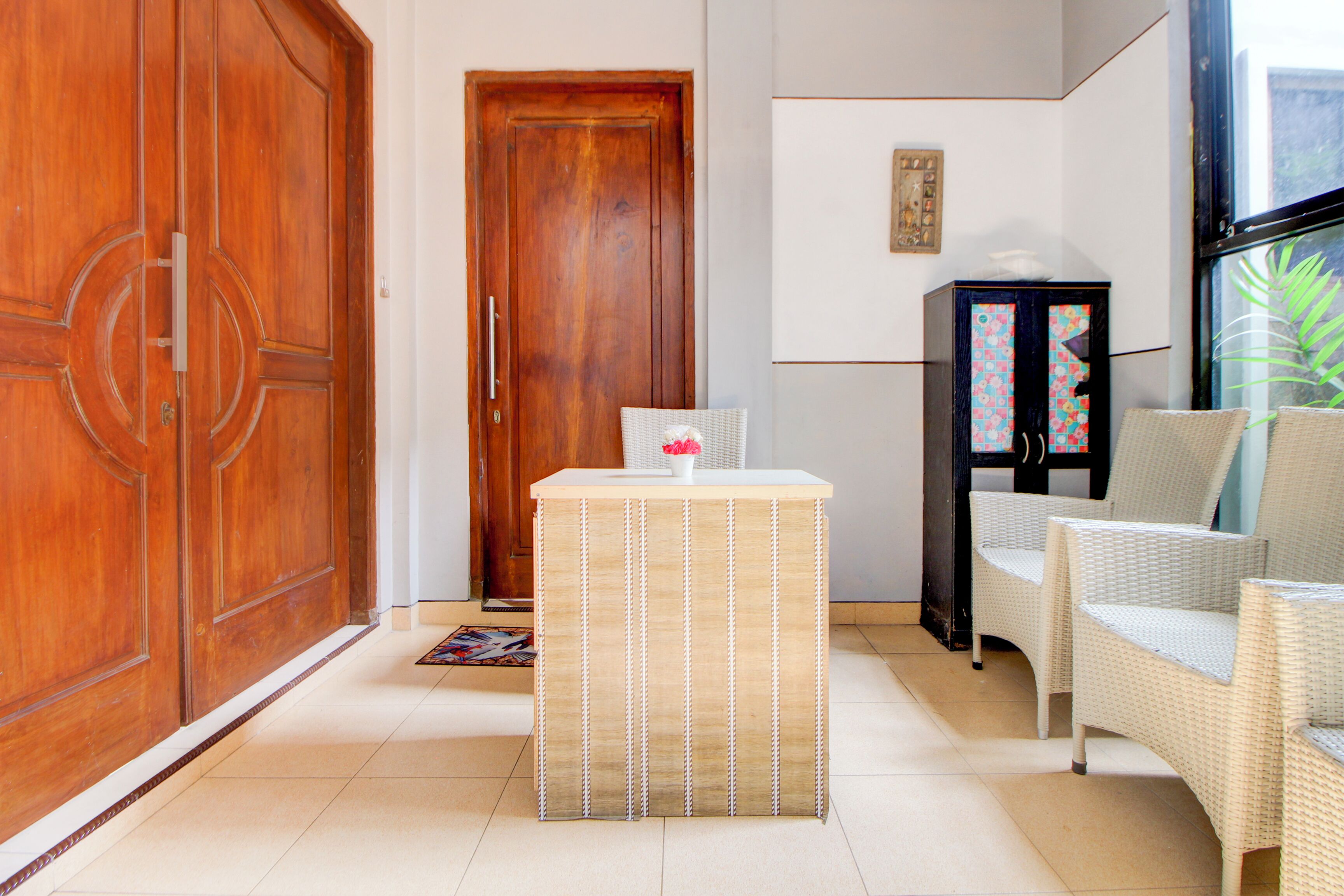 A room with a wooden door, a small table with a white vase, wicker chairs, and a cabinet with a colorful pattern.
