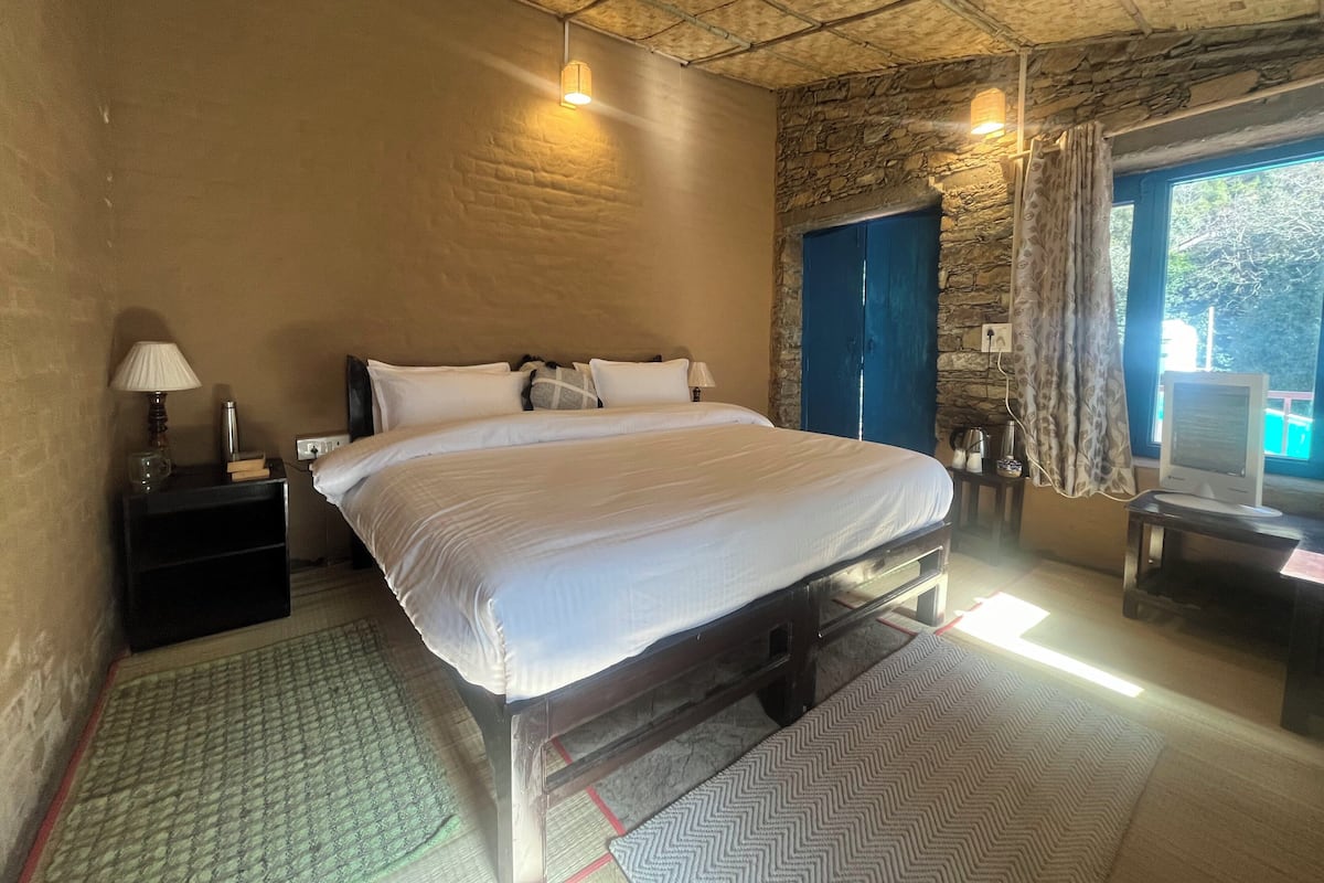 A bedroom with a large bed, a bedside table with a lamp, a small table with a computer, and a window with curtains.