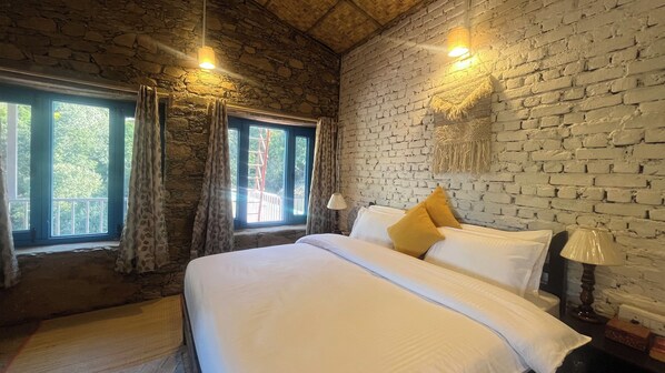 Un dormitorio con pared de piedra, una cama con ropa de cama blanca y un almohadón amarillo, dos lámparas de noche y una ventana con cortinas.