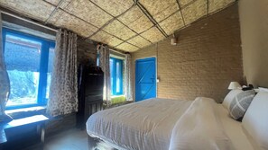 Un dormitorio con cama, ventana con cortinas, puerta azul y pared de ladrillo.