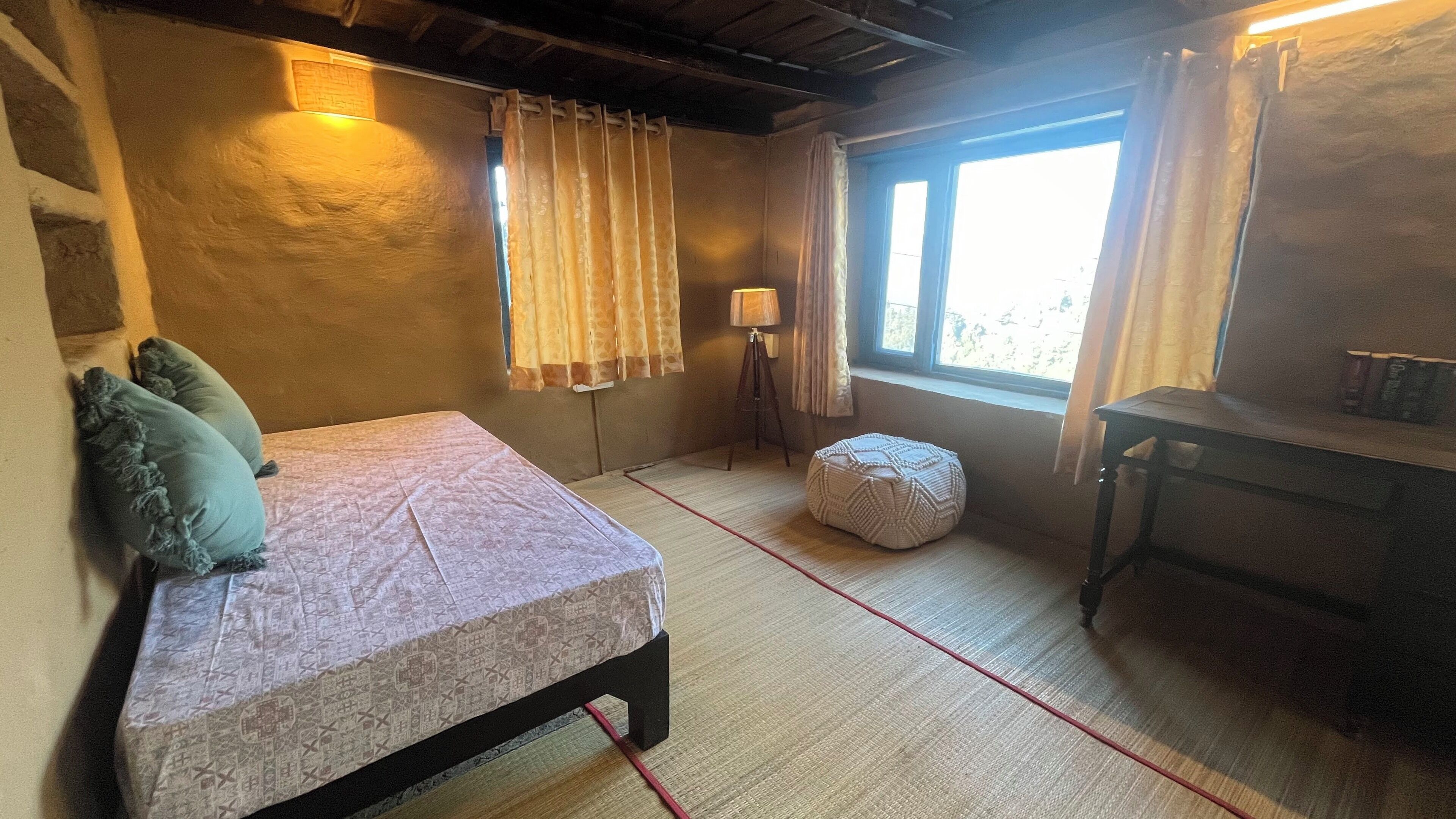 Quarto com cama, uma mesa pequena, luminária e janela com cortinas.