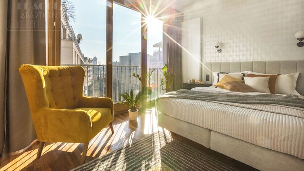 Ein modernes Schlafzimmer mit einem großen Bett, einem gelben Sessel und Blick auf die Stadt durch große Fenster.