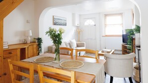 A dining area with a wooden table, chairs, and a vase with flowers. There is a staircase on the left and a door in the background.