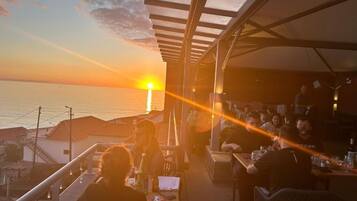 An outdoor dining area with tables and chairs, overlooking a sunset over the sea.
