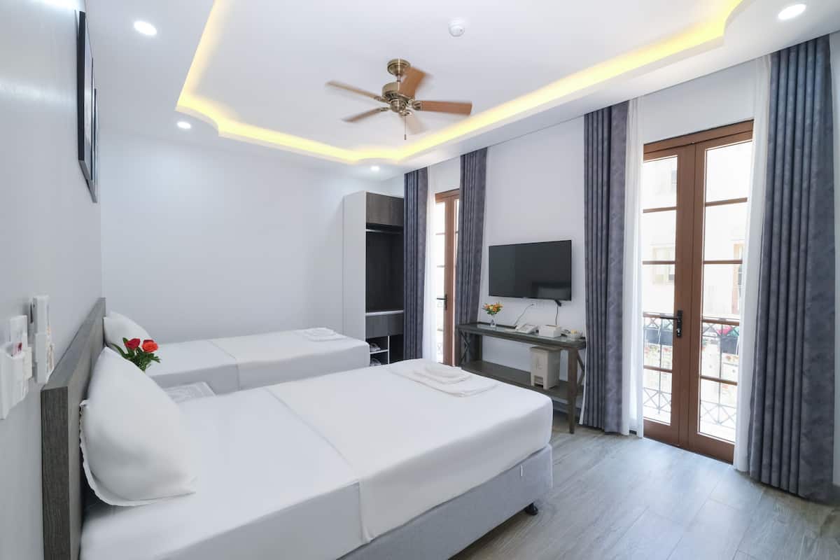 Une chambre d’hôtel moderne avec deux lits simples, un bureau doté d’une télévision à écran plat, un ventilateur de plafond et un balcon avec des rideaux.