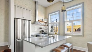 Een moderne keuken met een roestvrijstalen koelkast, een groot eiland met krukken, een hangende lamp en uitzicht door een raam.