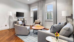 A modern living room with a sofa, armchair, and a coffee table. There is a flat-screen TV mounted on the wall, and a ceiling fan. - Perfect Location - King Street Condo! (Charleston)