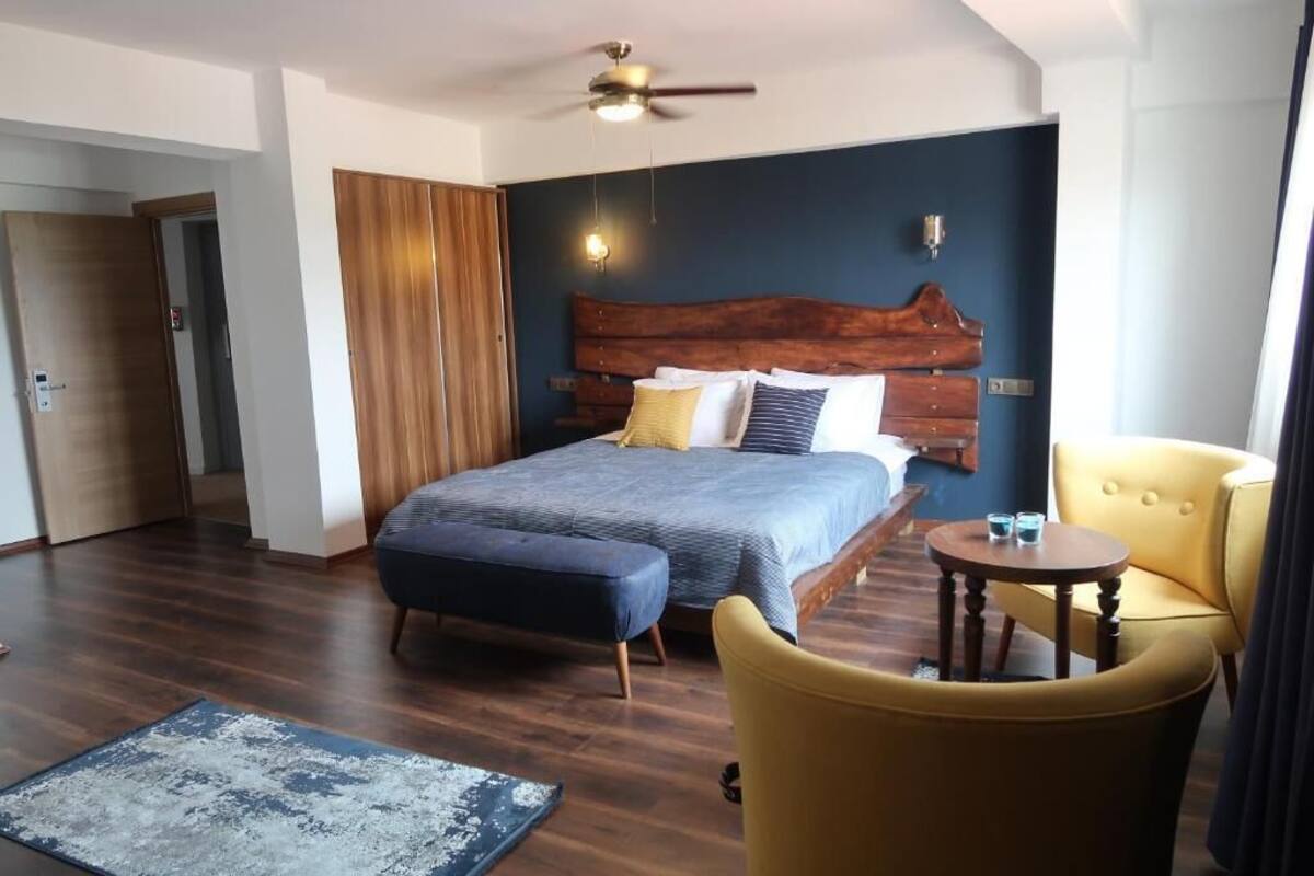 Une chambre d’hôtel avec un grand lit, une tête de lit en bois, une petite table ronde, un fauteuil jaune et un ventilateur de plafond.