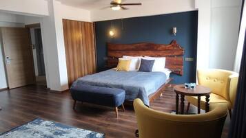 Camera d'hotel con un letto grande, una testiera in legno, un tavolino rotondo, una poltrona gialla e una ventola a soffitto.
