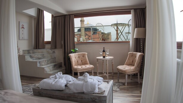 Ein Hotelzimmer mit Balkon, zwei Stühlen, einem kleinen Tisch mit Flasche und Gläsern, einer Treppe und Blick auf eine Moschee und einen Turm.