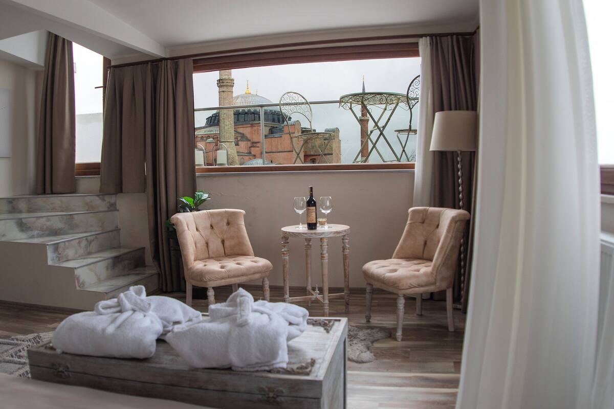 Une chambre d’hôtel avec un balcon, deux chaises, une petite table avec une bouteille et des verres, un escalier et une vue sur une mosquée et un clocher.