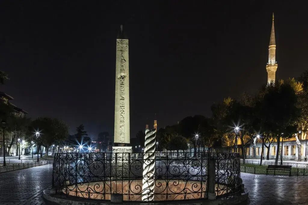 A monument with inscriptions, surrounded by a decorative fence, in a well-lit public square at night.