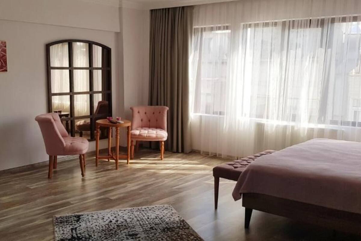 Une chambre d’hôtel avec un grand lit, deux fauteuils roses, une petite table, une grande fenêtre avec des rideaux et un tapis décoratif.