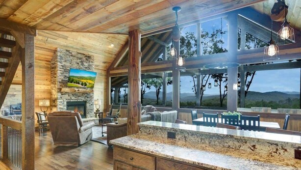 A spacious living area with a stone fireplace, wooden beams, and large windows offering a view of the outdoors. - Luxury Cabin w/ Stunning Mountain Views & Hot Tub (Blue Ridge)