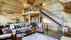 A spacious log cabin interior with a living area, a dining area, and a kitchen. A wooden staircase connects the two levels. - Luxury Cabin w/ Stunning Mountain Views & Hot Tub (Blue Ridge)
