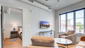 A modern living room with a sofa, armchair, and coffee table. There is a TV mounted on the wall and a small table with a lamp. The room has hardwood flooring and large windows. - Fire Tower | 1BR Hideaway in Downtown CHS with TV! (Charleston)
