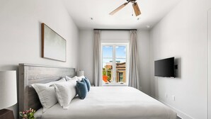 A modern hotel room with a large bed, a flat-screen TV mounted on the wall, a ceiling fan, and a window with curtains. - Fire Tower | Private 1BR King Suite + Rooftop View (Charleston)