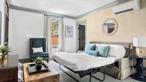 Una habitació d'hotel amb un llit, una cadira, una taula petita amb una planta, una taula de nit amb una làmpada i una unitat de aire condicionat a la paret.