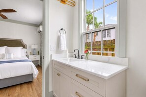 A hotel room with a bed, bedside table, lamp, and a window with a view of a wooden fence and palm trees. - Grand 9BR Charleston Downtown Home + Gym (Charleston)