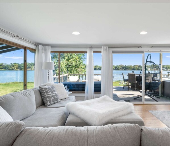 A modern living room with a large sofa, a white furry throw, and a view of a lake and deck area.