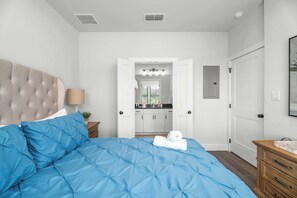 Un dormitorio con cama, mesita de noche, cómoda, una puerta y un baño visible a través de otra puerta abierta.