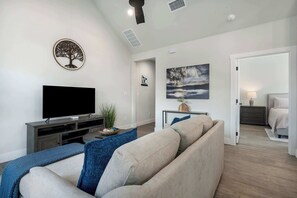 Ein Wohnzimmer mit einer Couch, einem Flachbildfernseher, Wandkunst mit einem Baumotiv, einem Gemälde und einer Tür, die in ein Schlafzimmer führt.