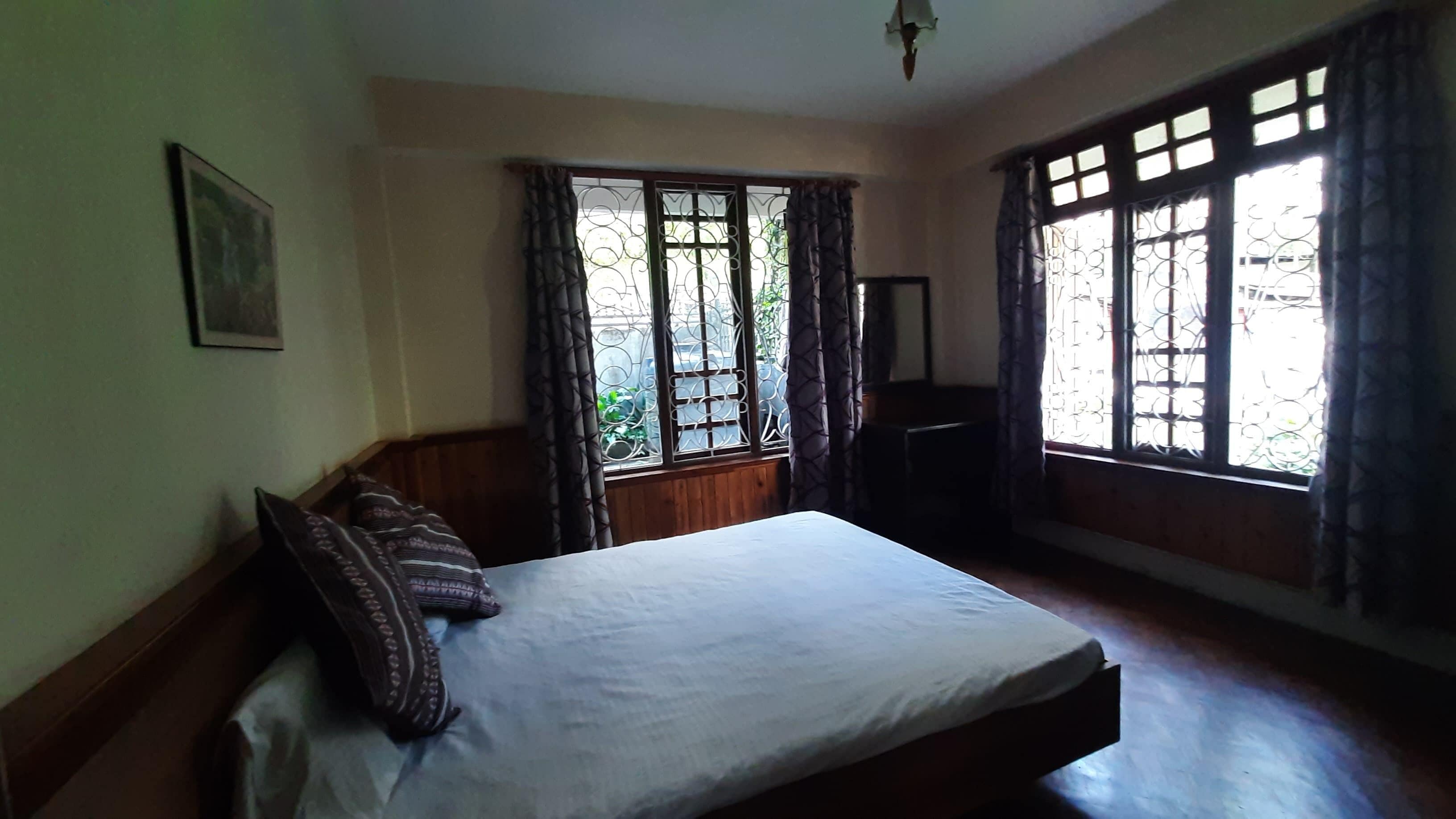 A bedroom with a bed, wooden headboard, a framed picture on the wall, and large windows with curtains.