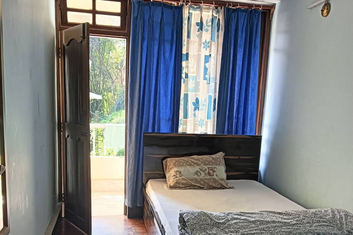 A bedroom with a bed, a wooden floor, a window with blue curtains, and a door leading outside.