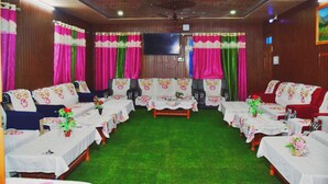 A room with a green carpet, wooden walls, and a chandelier. There are tables with white tablecloths and chairs with floral patterns. - Snow View Homestay (Sainj)